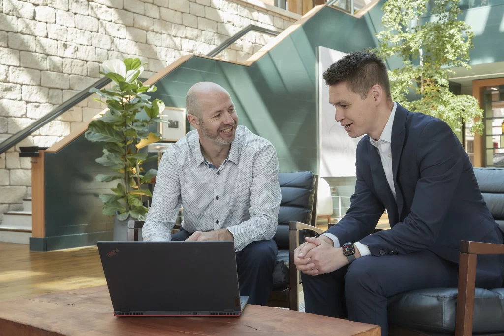 David Laws listening attentively while a client speaks and gestures, seated together at a laptop in an office atrium