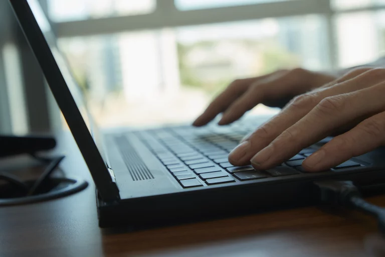 Close-up of hands typing on a laptop keyboard, office window light in the background